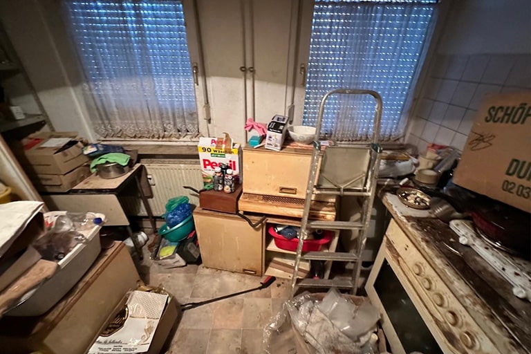 messi-v-10-min Eine überfüllte Küche mit überall verstreuten Kisten, Taschen und verschiedenen Gegenständen. Eine Leiter lehnt an einem Schrank. Im Hintergrund sind zwei Fenster mit transparenten Vorhängen zu sehen. Auf der rechten Seite ist ein alter Herd zu sehen. Reinigungsmittel und eine Packung Persil-Waschmittel liegen inmitten des Durcheinanders. Messie Küche entrümpeln und reinigen.